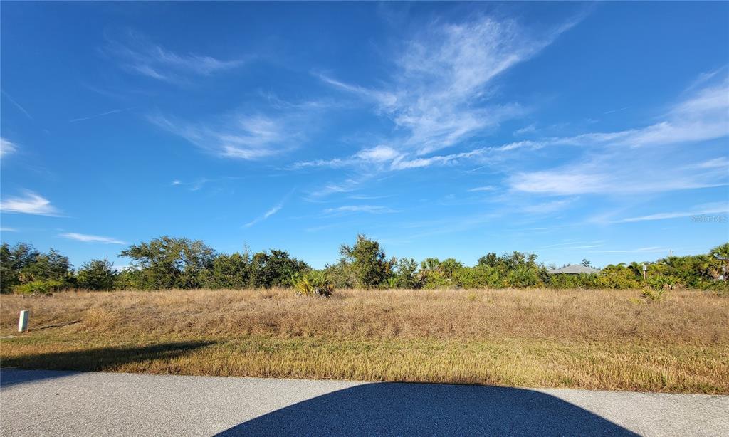 A vacant, grassy lot is viewed from an adjacent paved road under a blue sky — 13574 ALLAMANDA CIRCLE, PORT CHARLOTTE, FL