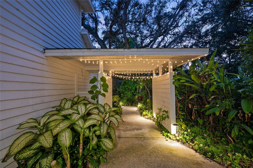 An exterior walkway features a covered patio with string lights and lush tropical landscaping — 6704 ARBOR OAKS DRIVE, BRADENTON, FL