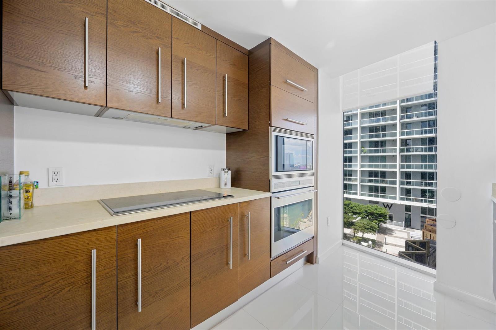 A modern kitchen features wood-grain cabinets, a flat cooktop, built-in appliances, and a large window — 475 Brickell Avenue # 1815, Miami FL 33131, MIAMI, FL