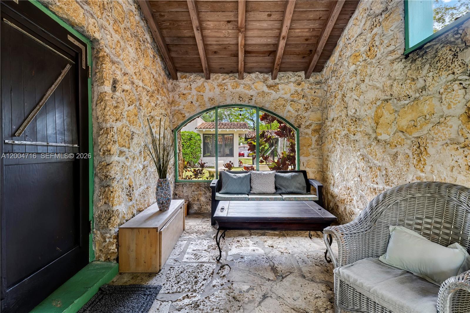 A rustic stone-walled sunroom features a wooden beamed ceiling, flagstone flooring, and a large window — 1301 Milan Ave, Coral Gables FL 33134, CORAL GABLES, FL