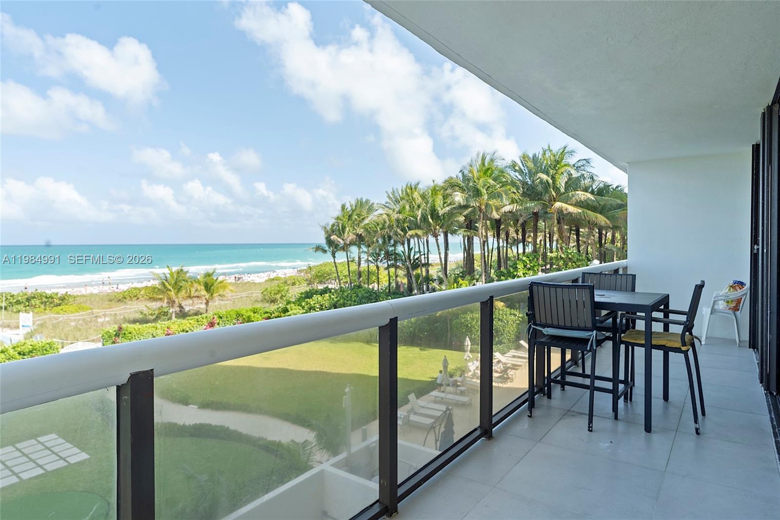 A balcony with outdoor furniture overlooks a grassy lawn, palm trees, and the ocean — 9455 Collins Ave # 409, Surfside FL 33154, SURFSIDE, FL