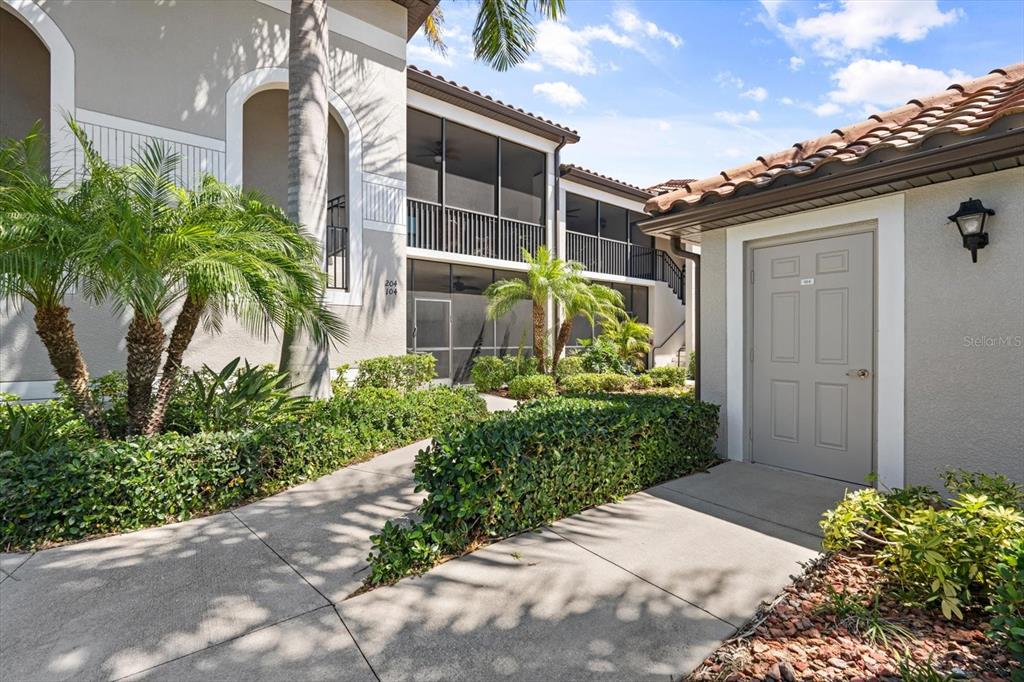 A concrete walkway leads to a gray door and a two-story building with screened balconies — 6705 GRAND ESTUARY TRAIL UNIT 104, BRADENTON, FL