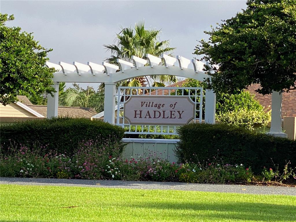 A white pergola structure stands behind a sign reading Village of Hadley amidst landscaping — 2530 SAFFRON LANE, THE VILLAGES, FL