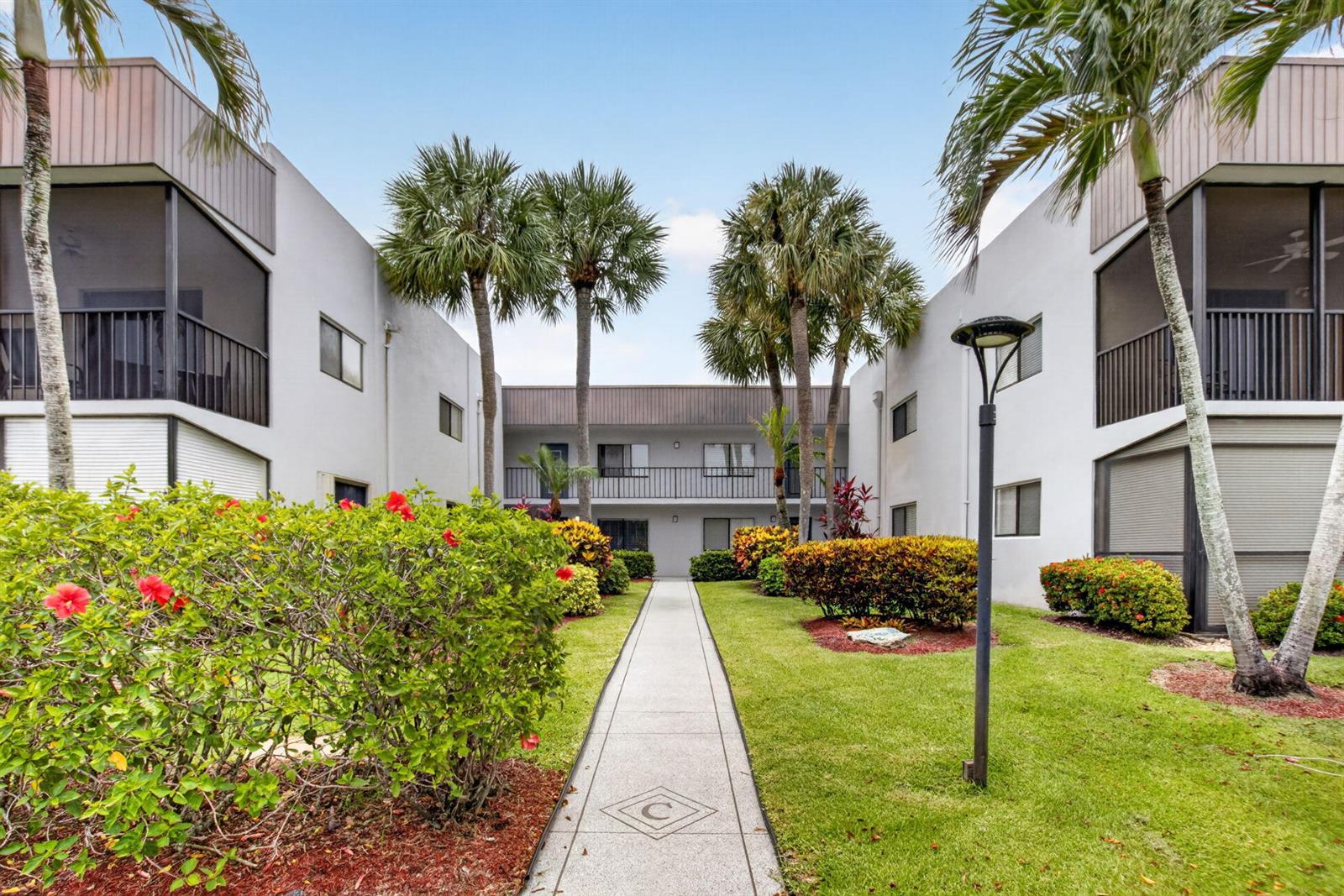 A concrete walkway leads between two white apartment buildings with palm trees and landscaping — 15054 Ashland Way # 82, Delray Beach FL 33484, DELRAY BEACH, FL