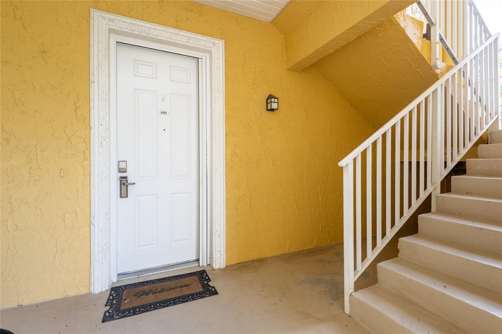 A white front door with a welcome mat sits beside an outdoor staircase — 6714 DALI AVENUE UNIT 101, LAND O LAKES, FL