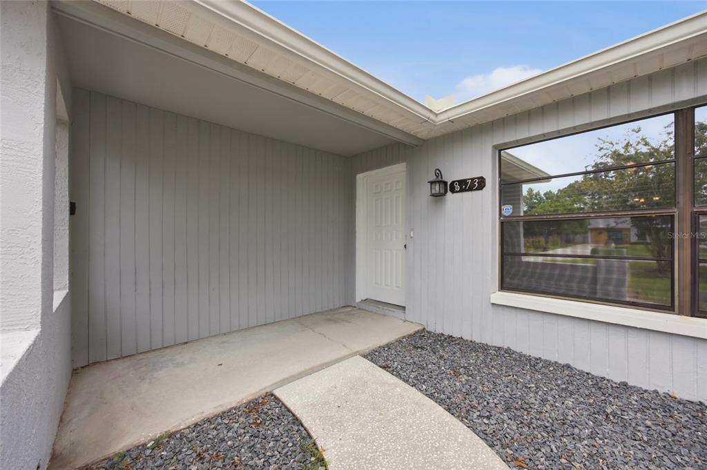 A gray-sided house exterior features a concrete walkway, a white front door, and a window — 8373 CAMPHOR DRIVE, SPRING HILL, FL