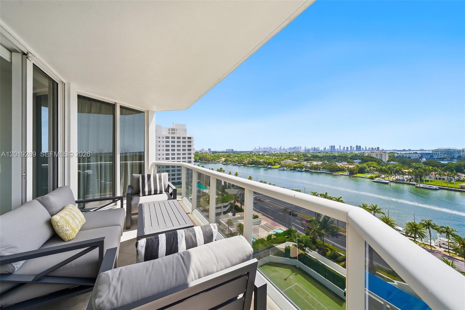 A balcony with outdoor seating overlooks a waterway, a road, and a distant city skyline — 4779 Collins Ave # 1206, Miami Beach FL 33140, MIAMI BEACH, FL