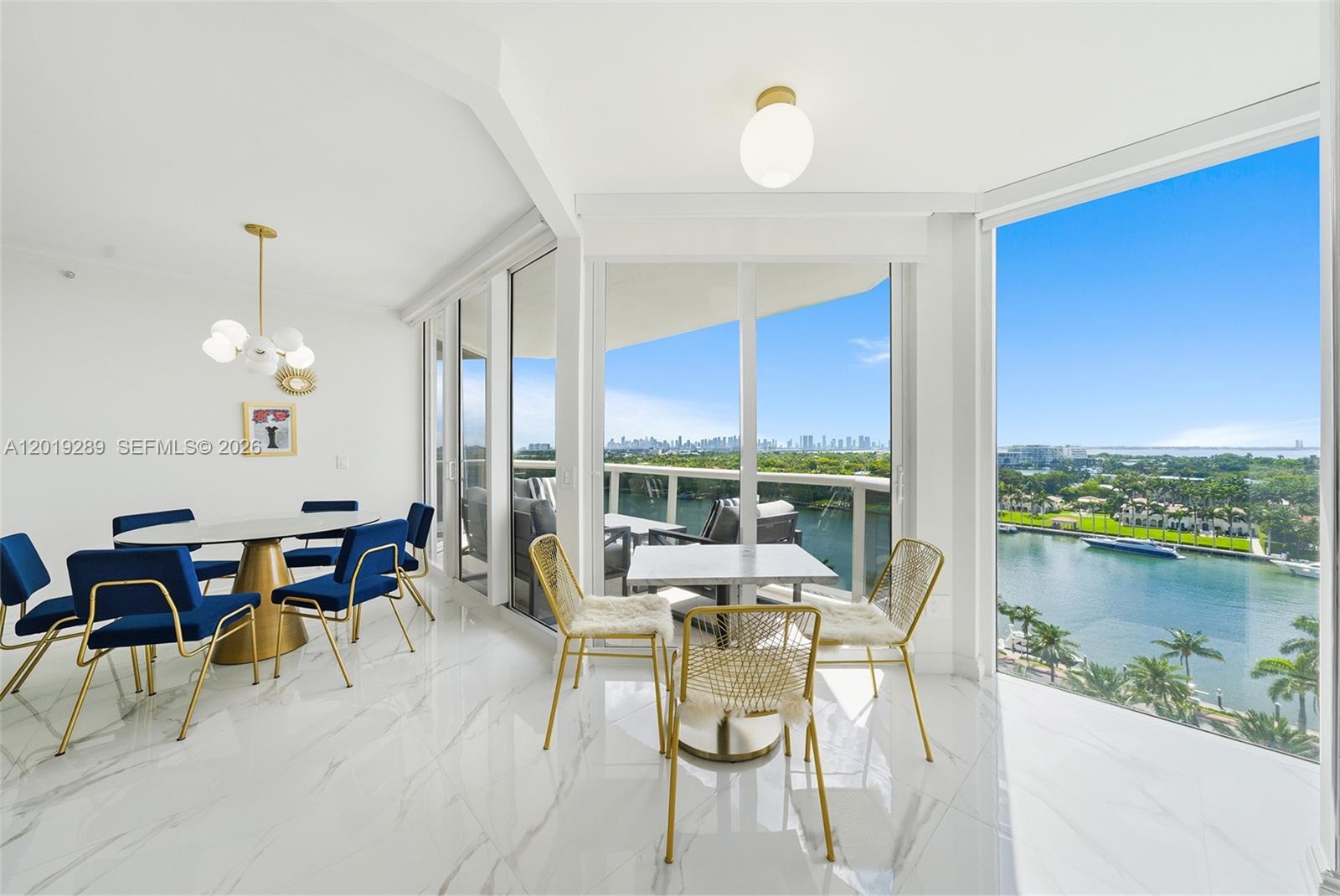A bright dining area features white marble floors, blue chairs, and floor-to-ceiling windows overlooking water — 4779 Collins Ave # 1206, Miami Beach FL 33140, MIAMI BEACH, FL