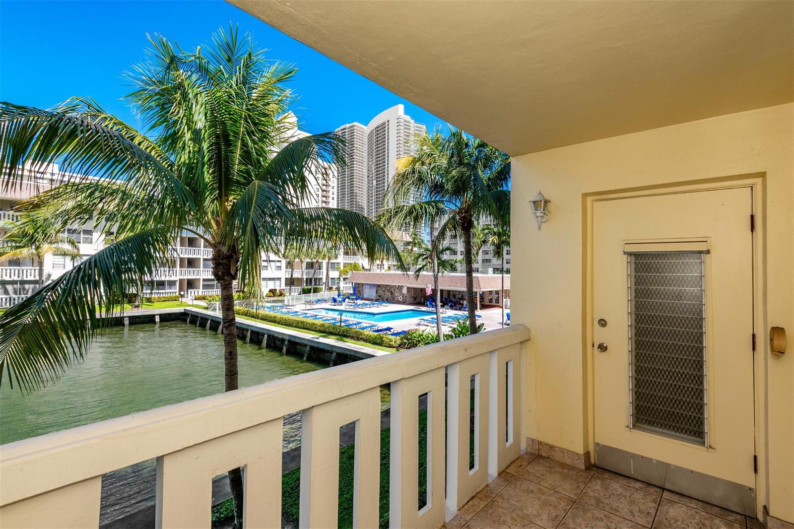 A balcony with a tiled floor overlooks a swimming pool, a canal, and palm trees — 1891 S Ocean Drive # 207, Hallandale Beach FL 33009, HALLANDALE BEACH, FL