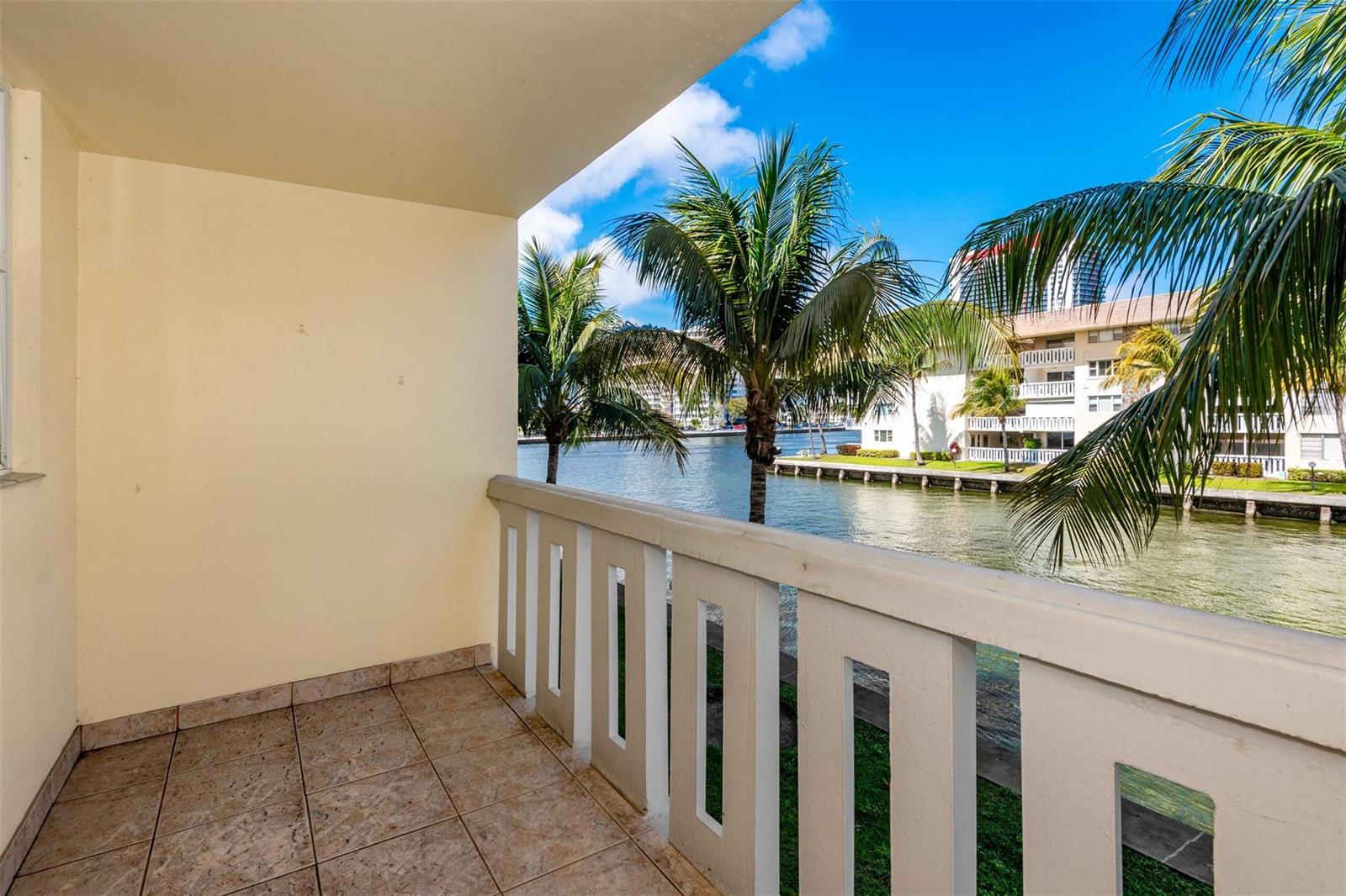 A tiled balcony overlooks a canal with palm trees and residential buildings in the background — 1891 S Ocean Drive # 207, Hallandale Beach FL 33009, HALLANDALE BEACH, FL
