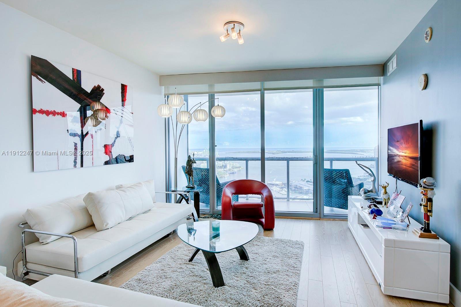 A modern living room features a white sofa, red chair, and floor-to-ceiling windows overlooking water — 888 Biscayne Blvd # 4007, Miami FL 33132, MIAMI, FL