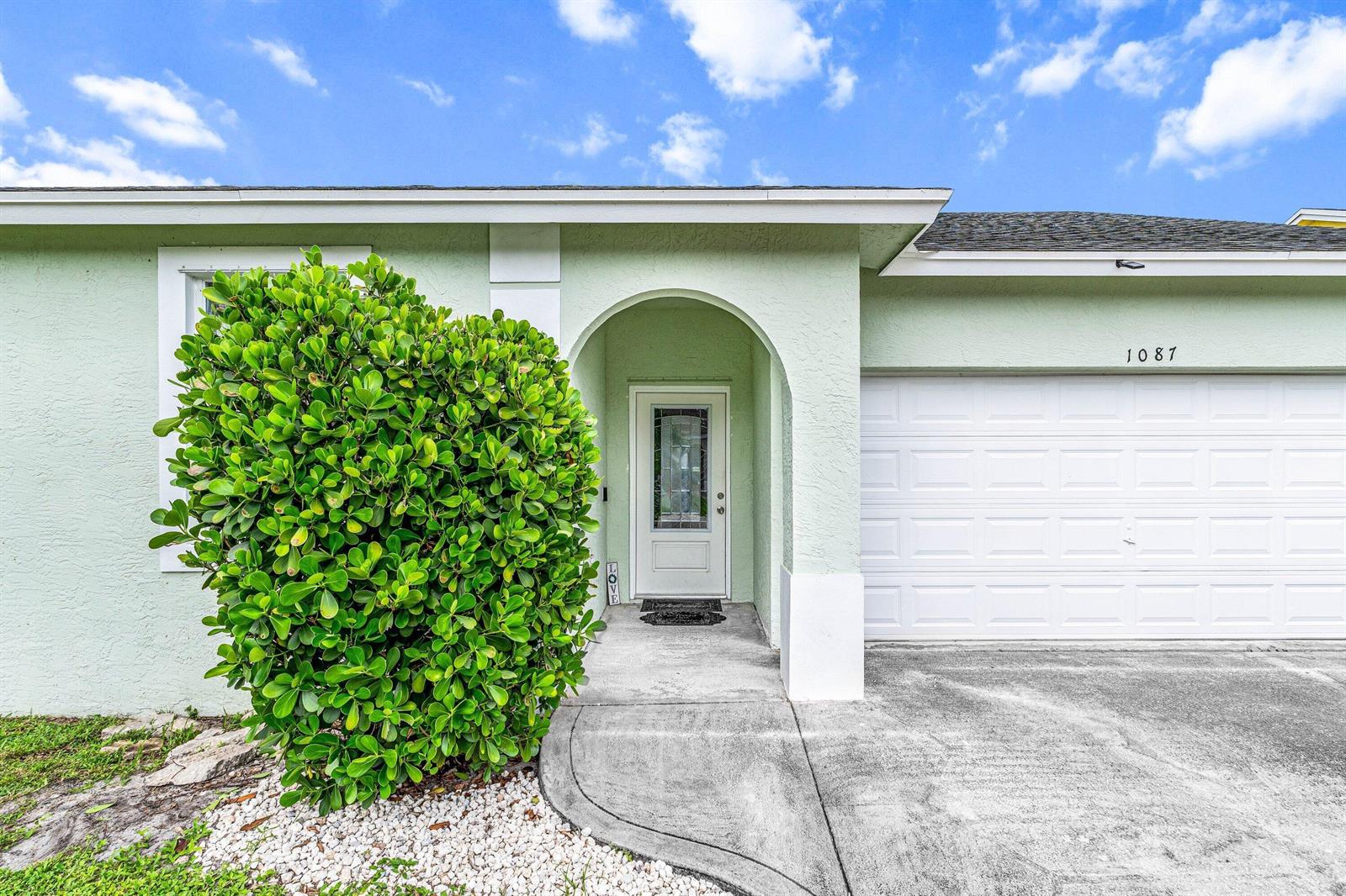 A light green house exterior features a white garage door, arched entryway, and large bush — 1087 Salmon Isle, FL 33413, FL
