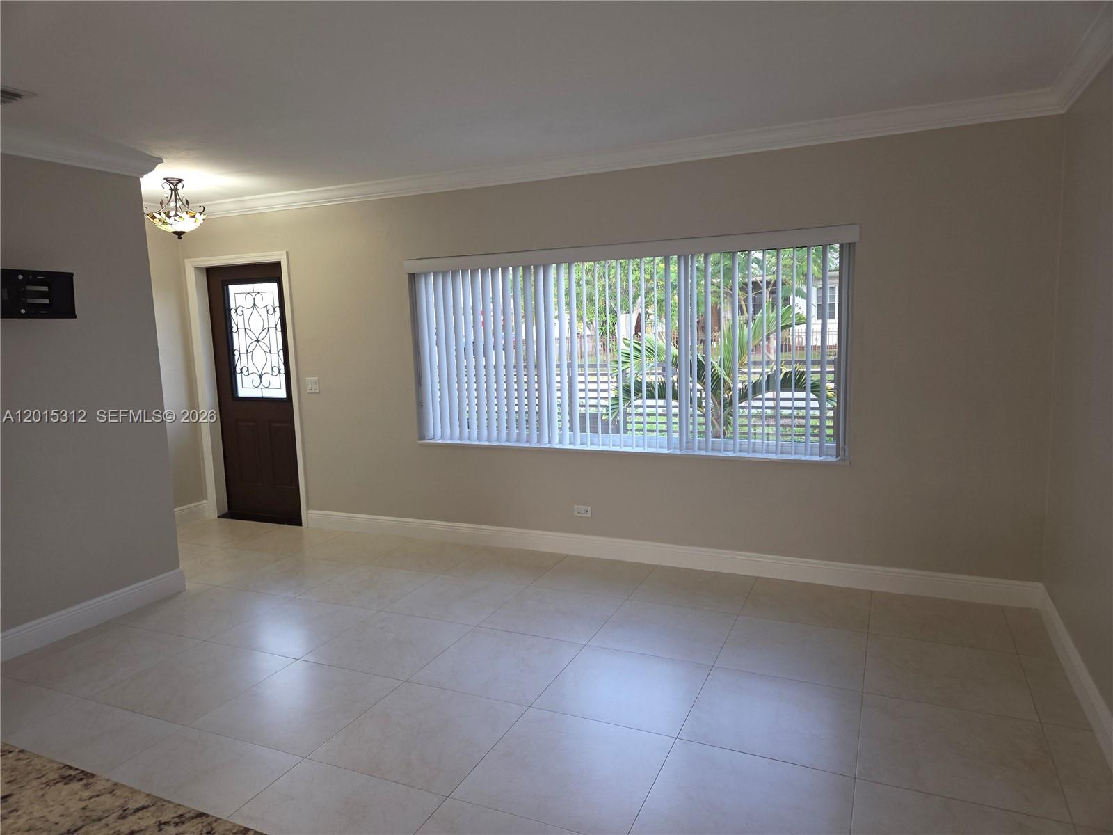 An empty room features light-colored tile flooring, beige walls, a large window with vertical blinds, and a dark front door — 1680 E 5th Ave # 1690, Hialeah FL 33010, HIALEAH, FL