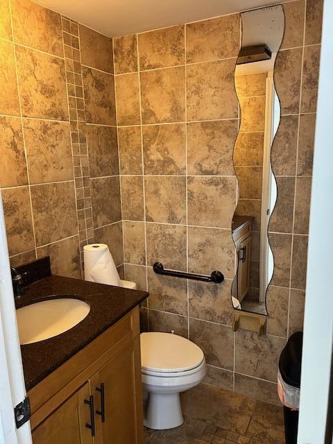 A small bathroom features tan stone-look wall tiles, a dark vanity, a toilet, and a wavy mirror — 1775 Blount Road # 409, Pompano Beach FL 33069, POMPANO BEACH, FL