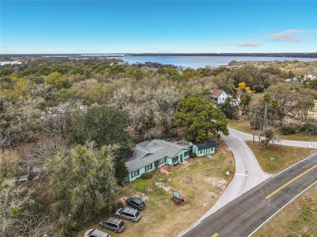 An aerial view shows a single-story house with a green roof surrounded by trees near water — 6942 OLD HIGHWAY 441 S, MOUNT DORA, FL