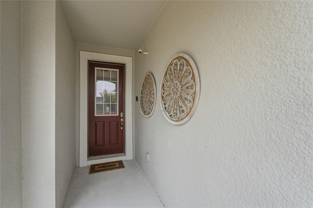 A covered entryway features a maroon front door, two circular wall decorations, and a welcome mat — 9083 SUNSHINE RIDGE LOOP, KISSIMMEE, FL