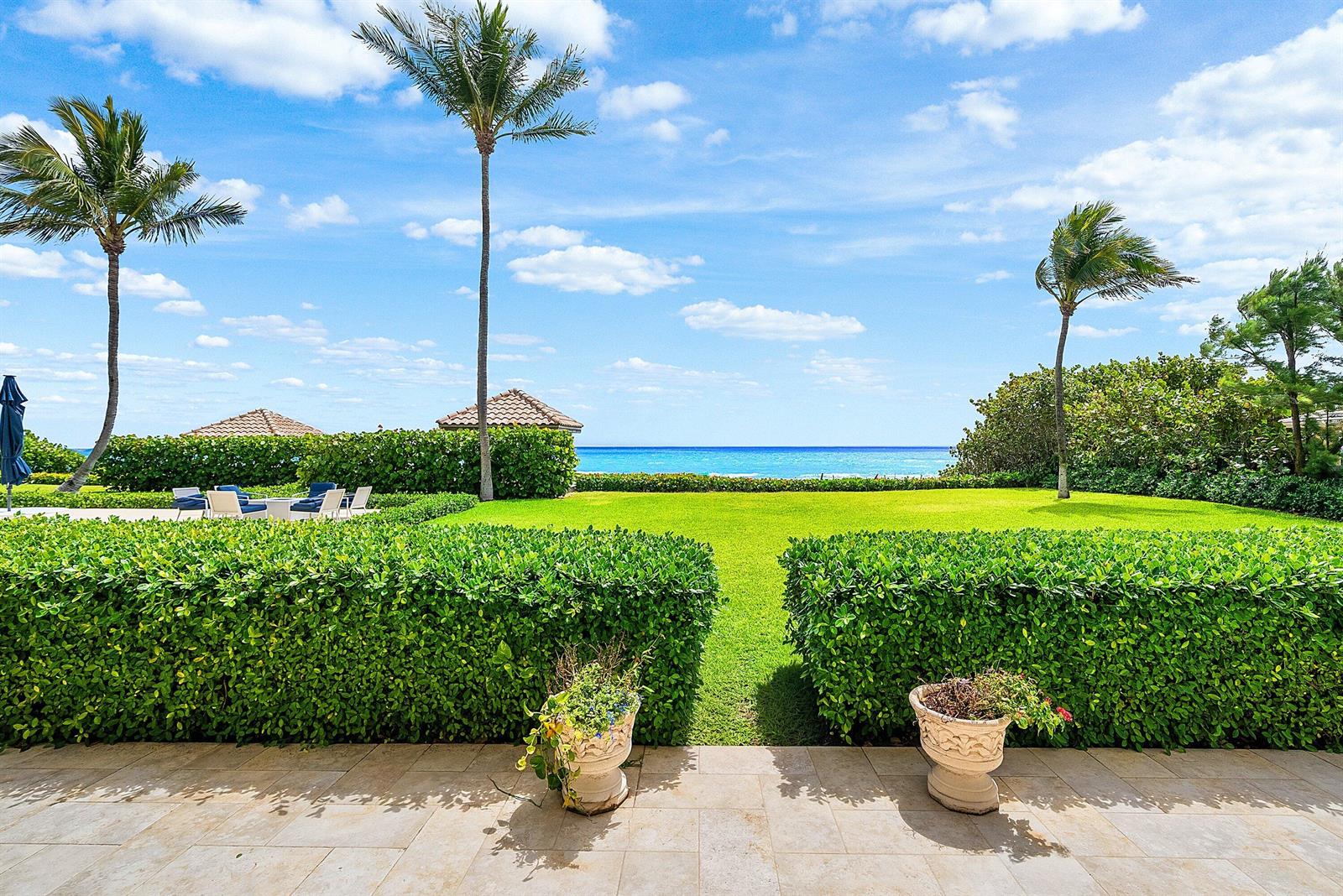 A stone patio overlooks a manicured lawn, trimmed hedges, palm trees, and the ocean — 3951 N Ocean Boulevard # 101, Gulf Stream FL 33483, GULF STREAM, FL