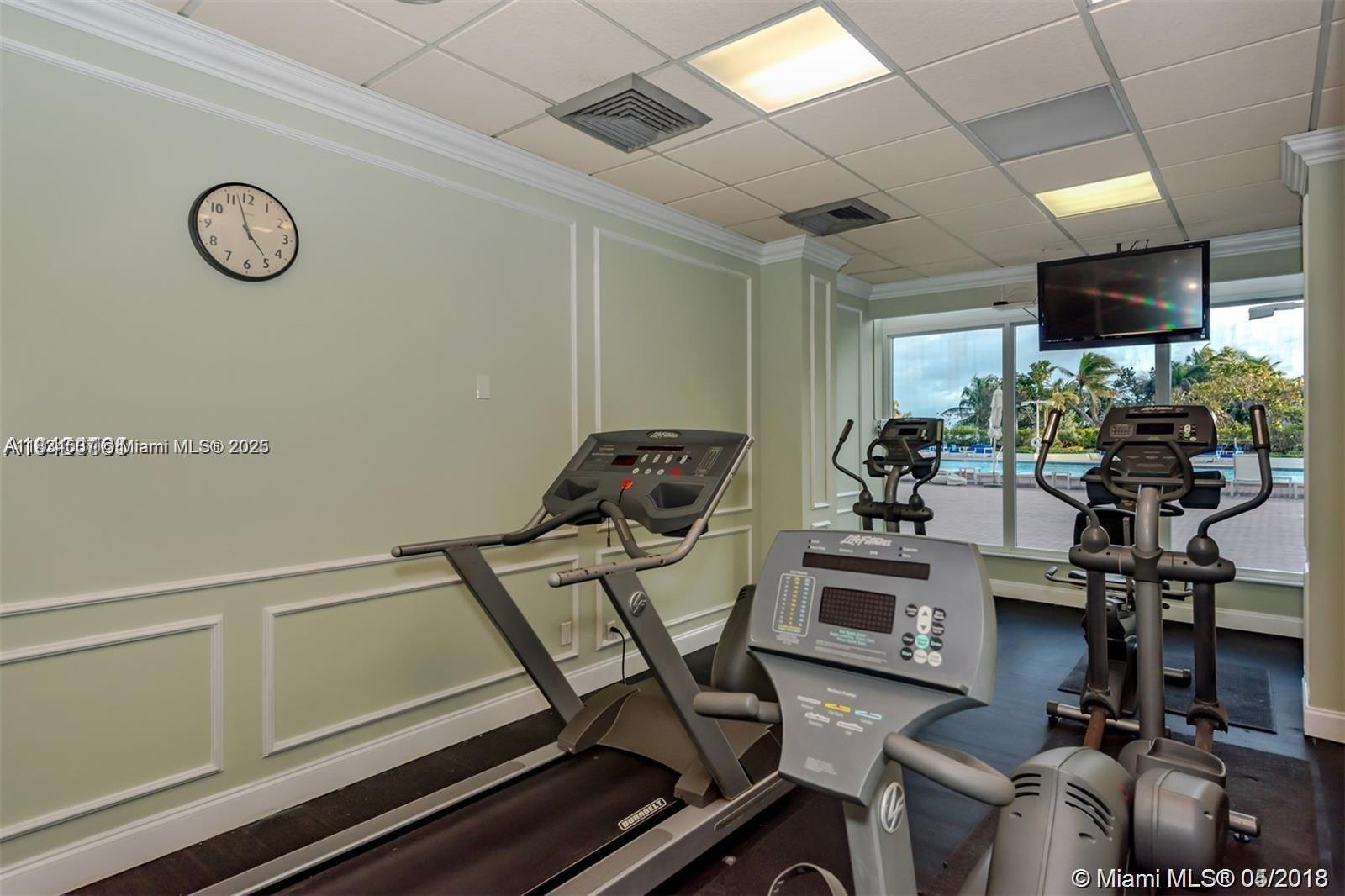 A gym features treadmills and elliptical machines facing a window with a pool view — 3180 S Ocean Dr # 1602, Hallandale Beach FL 33009, HALLANDALE BEACH, FL