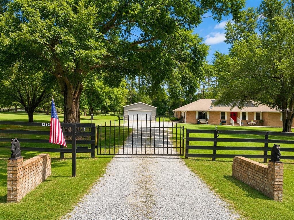 A gravel driveway leads through a black metal gate to a brick house and garage — 12820 NW 116TH PLACE, ALACHUA, FL