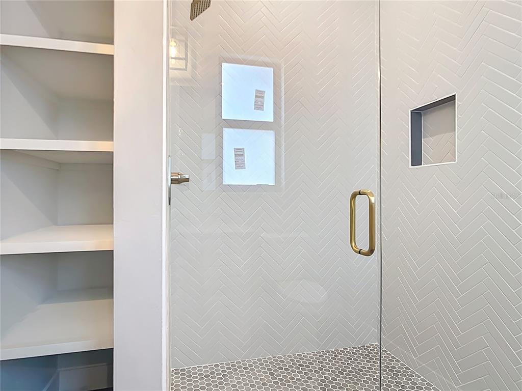 A glass-enclosed shower with herringbone wall tiles and a hexagonal tile floor next to shelving — 7152 BOUGAINVILLEA STREET, ENGLEWOOD, FL