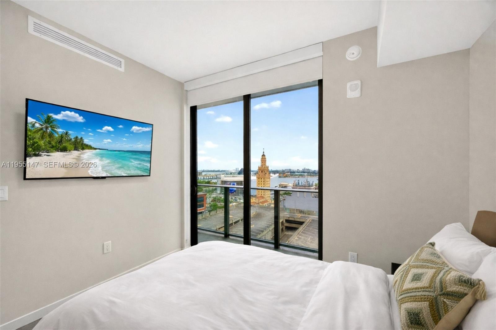 A bedroom features a wall-mounted television, a white bed, and a balcony with city views — 121 NE 5th Street # 1913, Miami FL 33132, MIAMI, FL