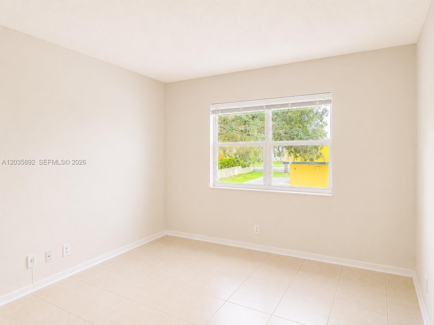 An empty bedroom features beige walls, light-colored tile flooring, and a window overlooking greenery — 280 S Cypress Rd # 310, Pompano Beach FL 33060, POMPANO BEACH, FL