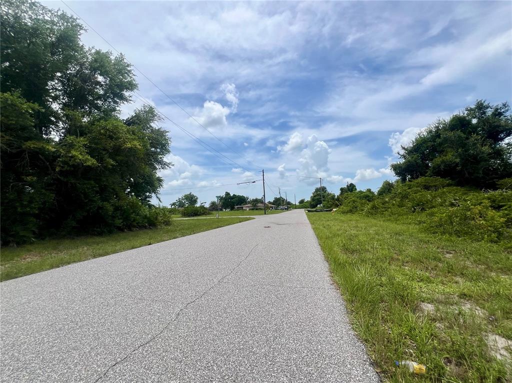 A paved road stretches through a grassy, undeveloped area with trees under a blue sky — 12322 ALBRECHT TERRACE, PORT CHARLOTTE, FL