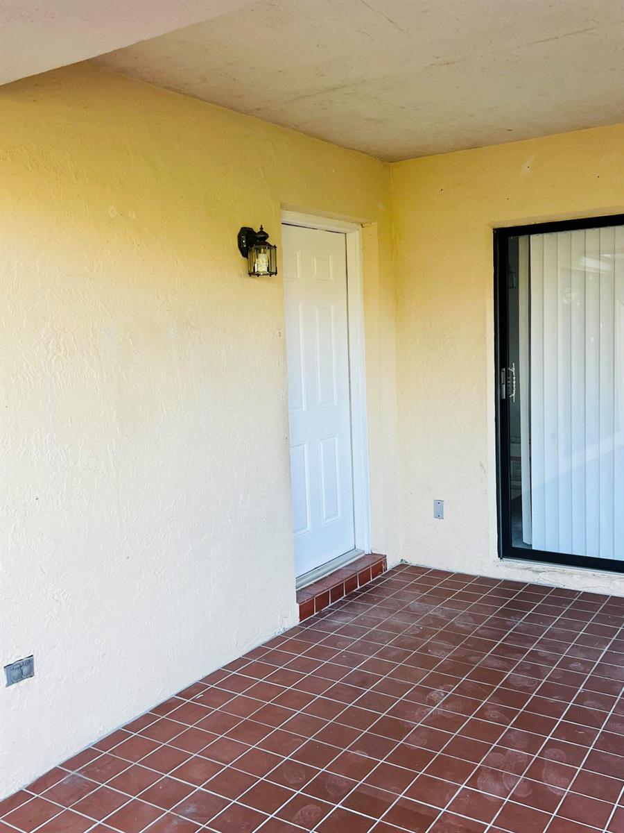 A covered patio features yellow stucco walls, a white door, and brown tiled flooring — 7482 E Courtyard Run E, Boca Raton FL 33433, BOCA RATON, FL