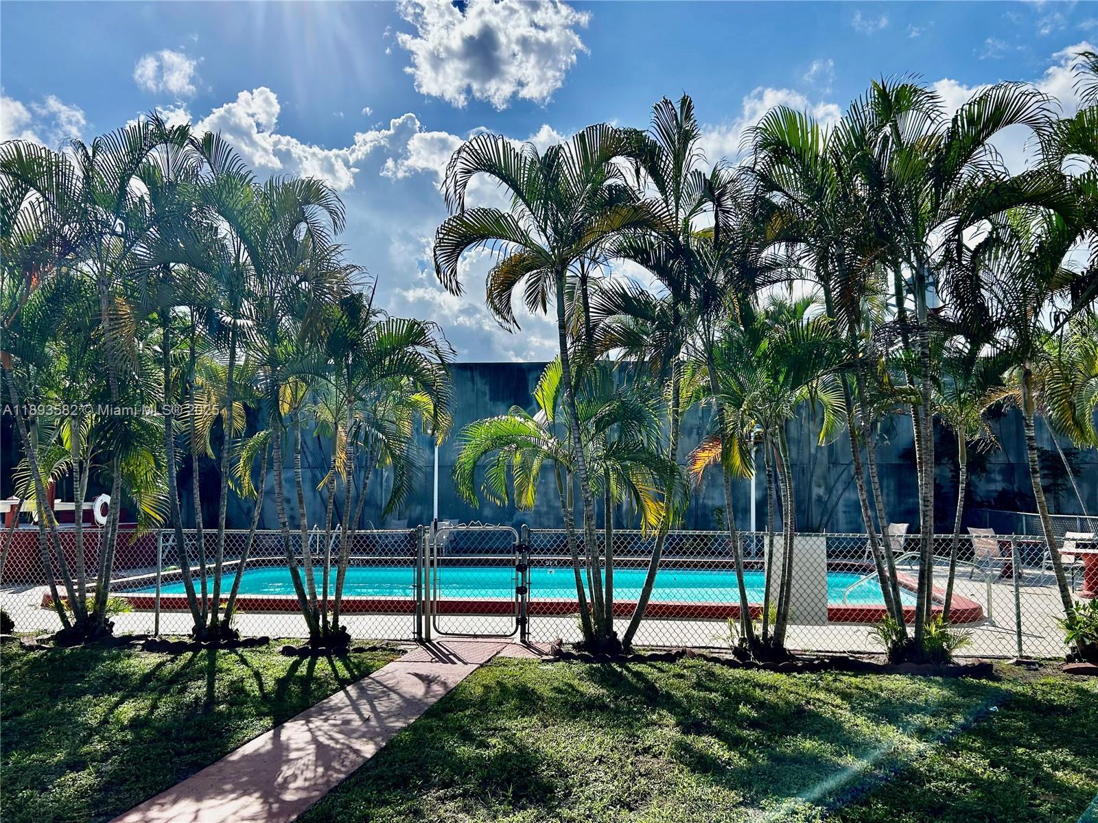 A fenced swimming pool surrounded by palm trees and a grassy lawn under a sunny sky — 2918 Funston St # 29B, Hollywood FL 33020, HOLLYWOOD, FL