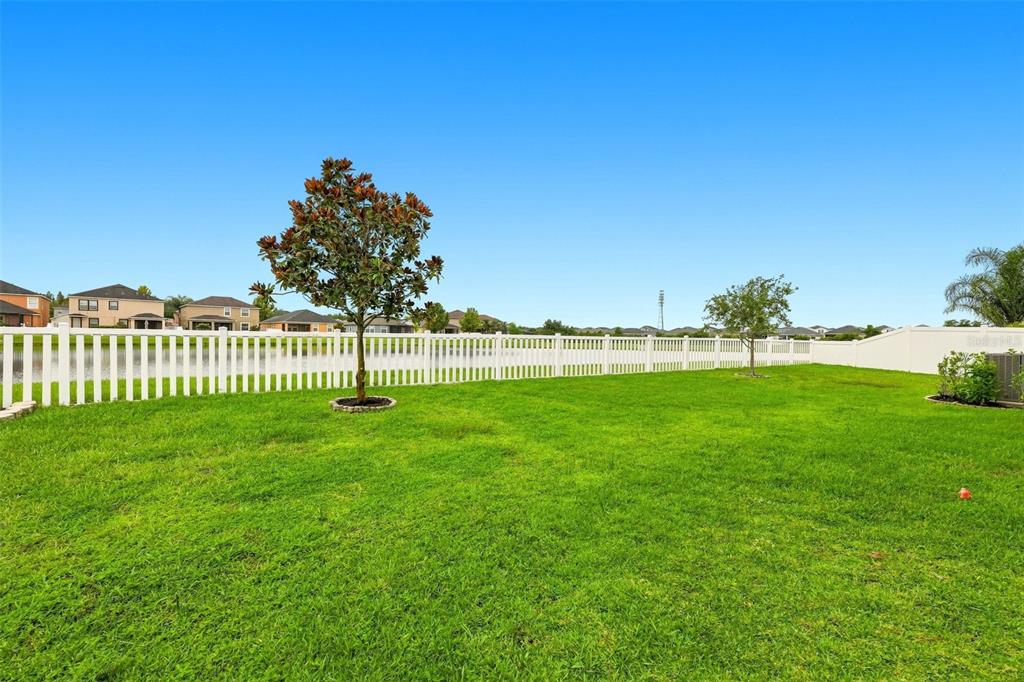 A grassy backyard features a white vinyl fence, two small trees, and a distant pond — 12335 BALLENTRAE FOREST DRIVE, RIVERVIEW, FL