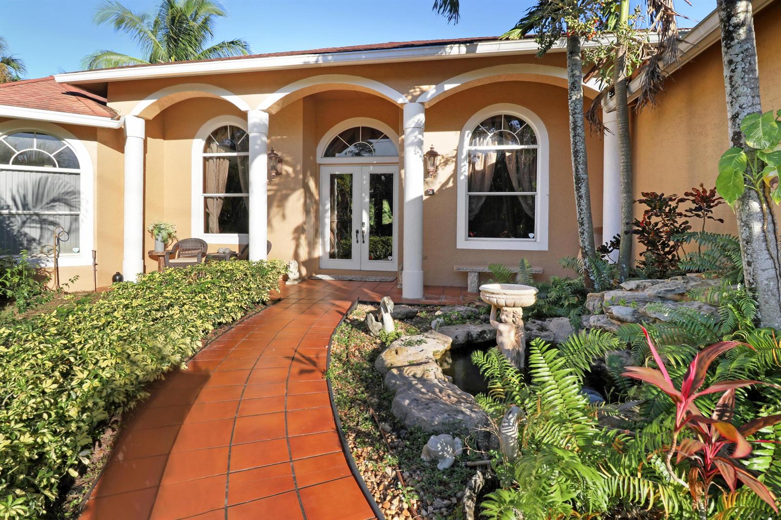 A terracotta walkway leads to the arched entryway of a tan house with a pond — 7086 Hall Boulevard, FL 33470, FL