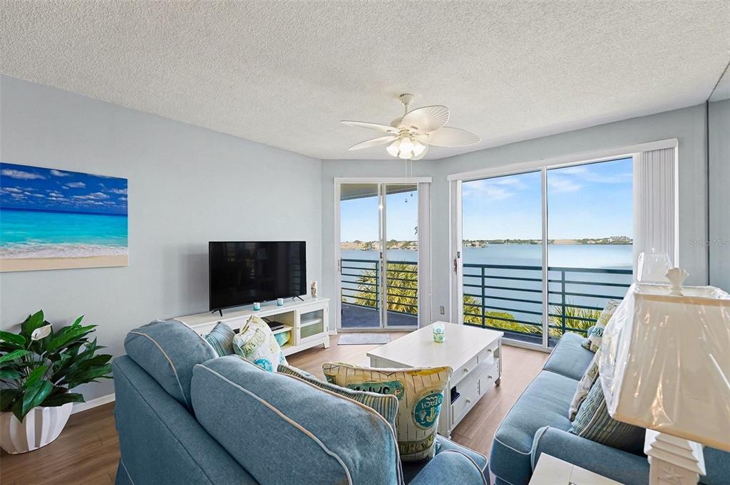 A living room features blue sofas, a television, and a balcony overlooking a large lake — 8021 SAILBOAT KEY BOULEVARD S UNIT 304, ST PETE BEACH, FL