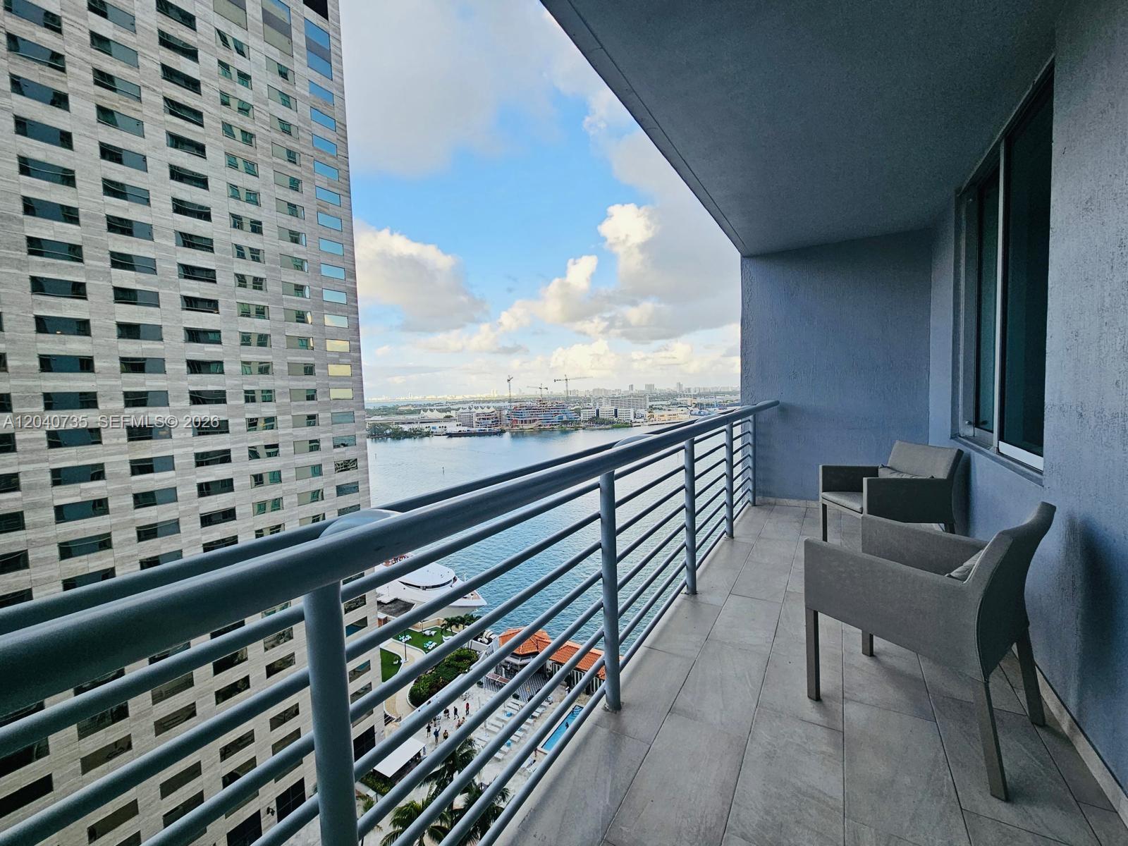 A balcony with two gray chairs overlooks a body of water and a tall building — 335 S Biscayne Blvd # 2202, Miami FL 33131, MIAMI, FL