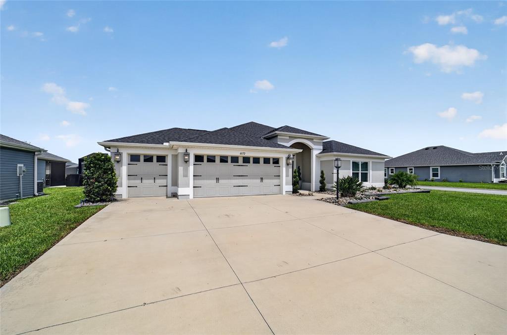 A single-story house with a three-car garage and a concrete driveway under a blue sky — 6172 DANIELSON LOOP, THE VILLAGES, FL