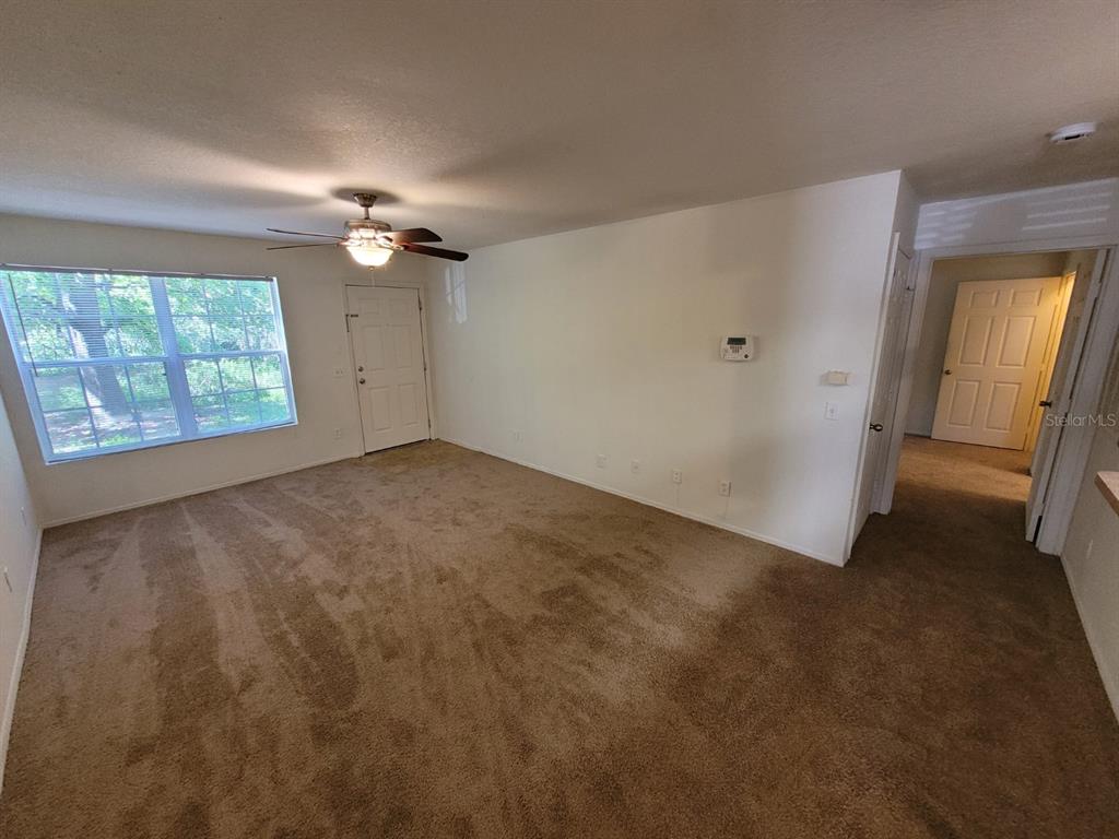 An empty living room features beige carpet, a ceiling fan, and a white front door — 36106 LAKE CHASE BOULEVARD UNIT 104, ZEPHYRHILLS, FL