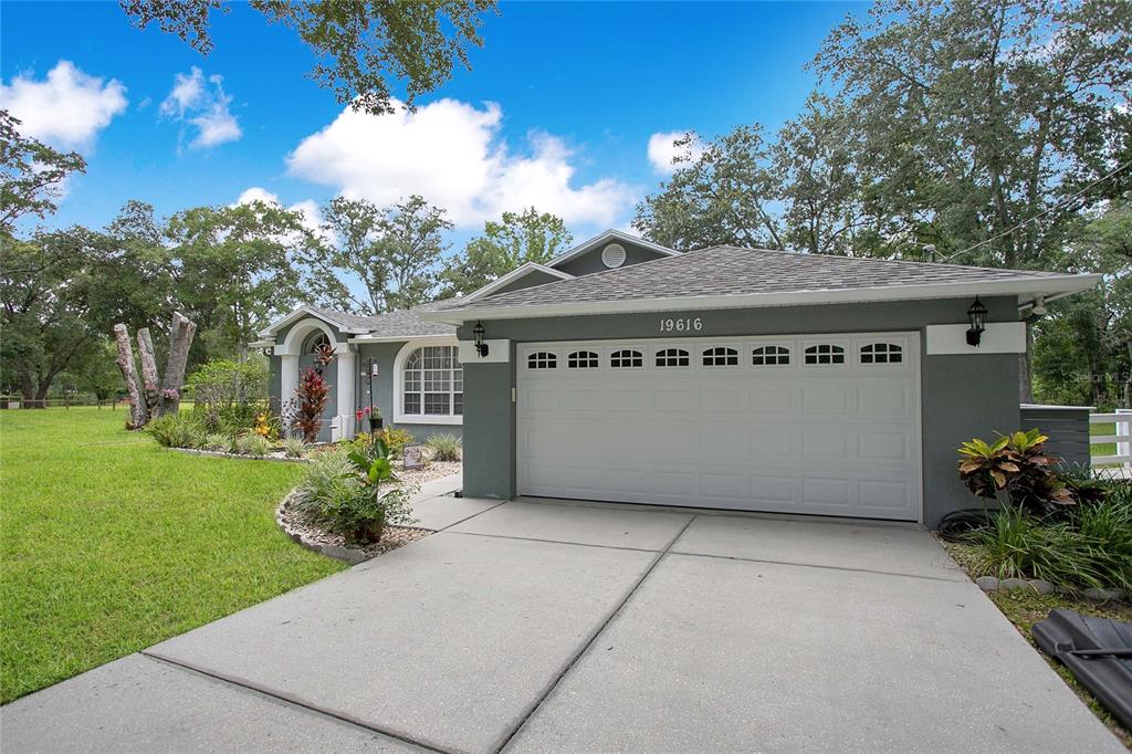 A gray single-story house features a white two-car garage door and a landscaped front yard — 19616 LIVINGSTON AVENUE, LUTZ, FL