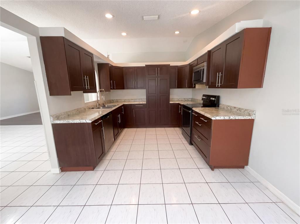 A U-shaped kitchen features dark wood cabinets, light stone countertops, and white tile flooring — 1750 EVERGREEN COURT, KISSIMMEE, FL