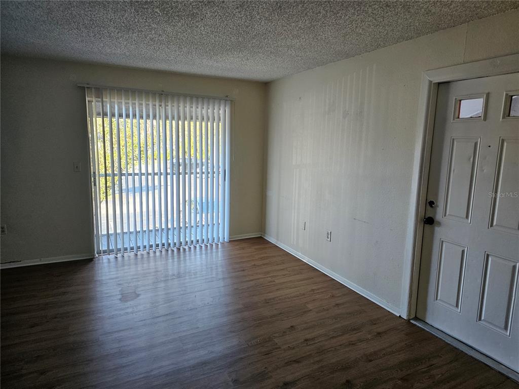 An empty room features wood-look flooring, a sliding glass door with vertical blinds, and a door — 5400 26TH STREET W UNIT E80, BRADENTON, FL