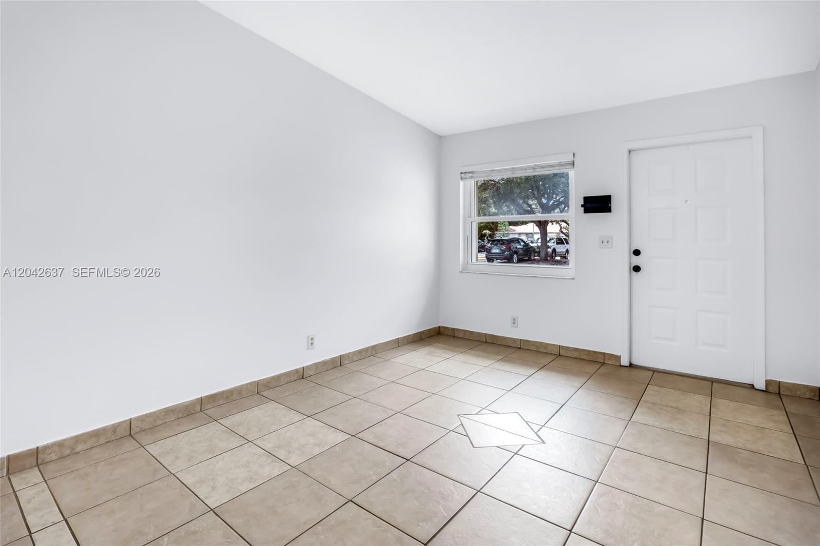 An empty room features white walls, tan tile flooring, a window, and a white door — 5020 Sanctuary Way # C, West Palm Beach FL 33417, WEST PALM BEACH, FL