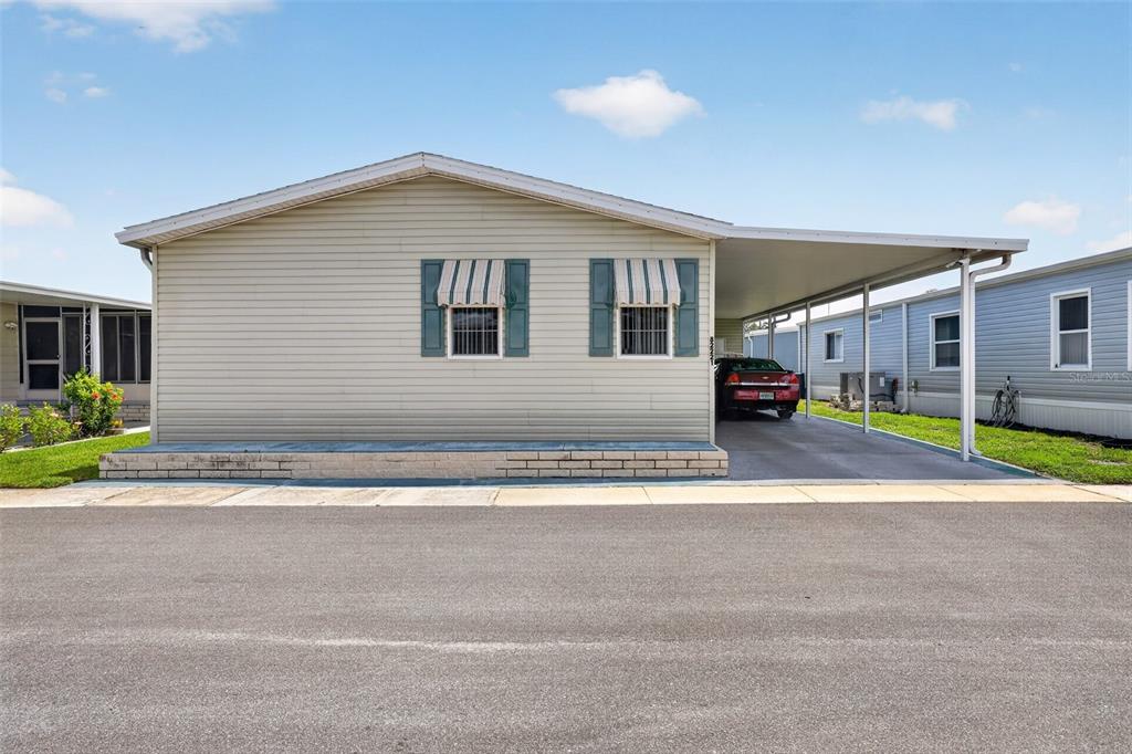 A single-story manufactured home with light siding, a carport, and a brick-bordered front patio — 82221 F STREET N UNIT 221, PINELLAS PARK, FL
