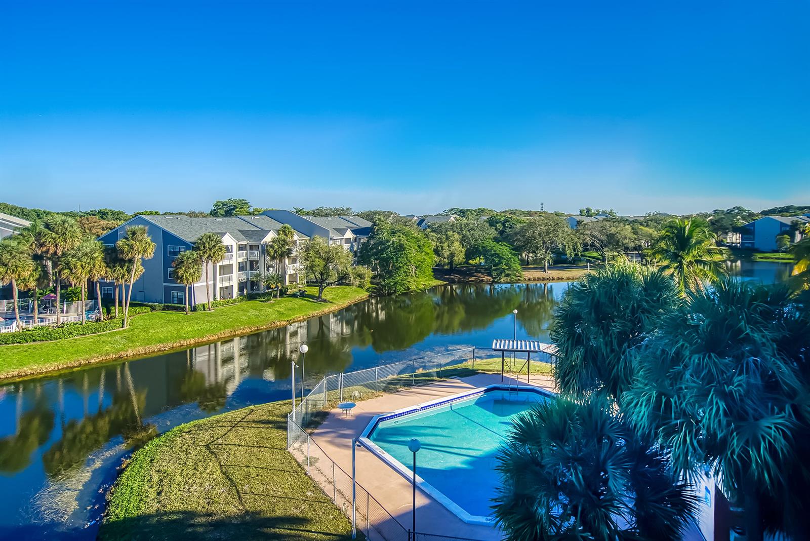 An outdoor swimming pool sits beside a lake with apartment buildings in the background — 8030 Hampton Boulevard # 502, North Lauderdale FL 33068, NORTH LAUDERDALE, FL