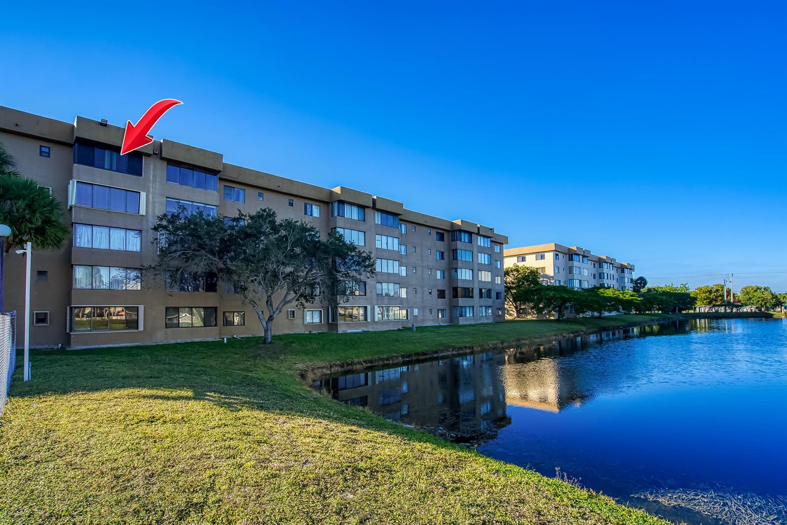 An exterior view of a multi-story apartment building beside a pond with a red arrow — 8030 Hampton Boulevard # 502, North Lauderdale FL 33068, NORTH LAUDERDALE, FL