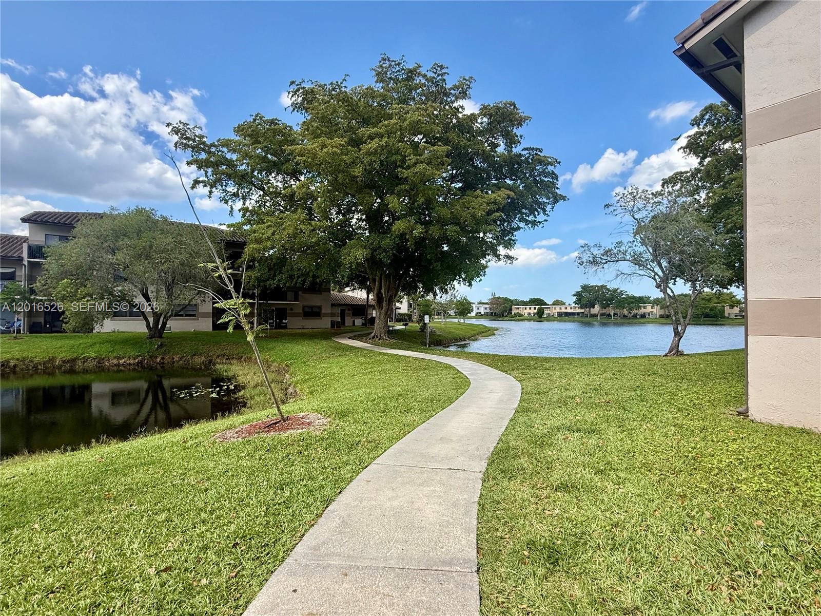 A concrete path winds through a grassy lawn between apartment buildings and a lake — 9827 Nob Hill Ct # 0, Sunrise FL 33351, SUNRISE, FL