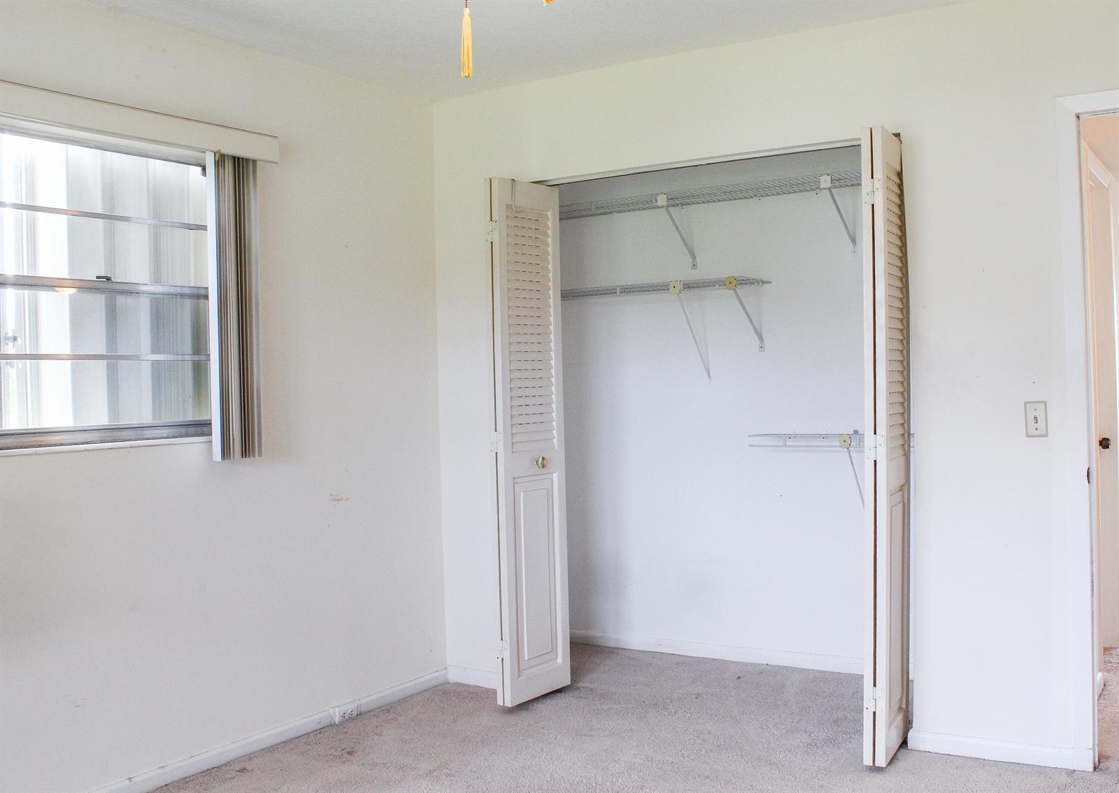 An empty bedroom with white walls, beige carpet, a window, and an open closet — 66 Brighton, Boca Raton FL 33434, BOCA RATON, FL