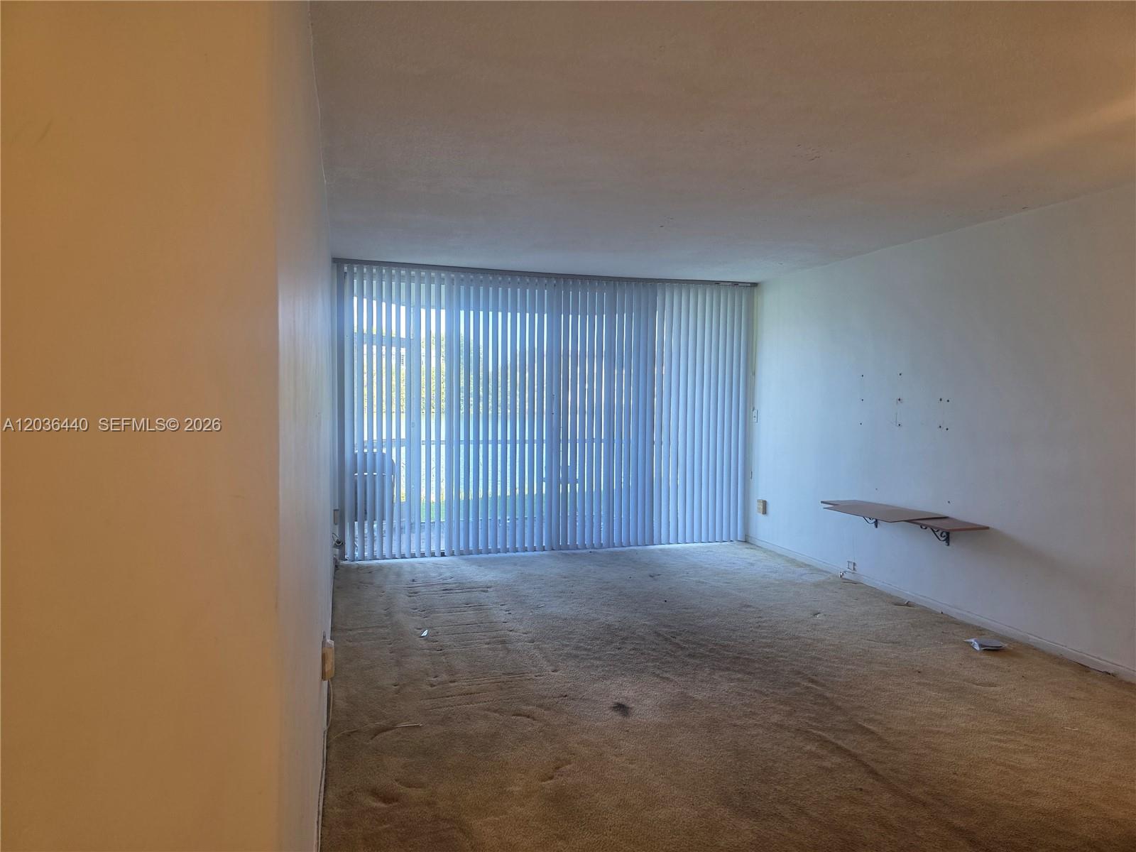 An empty room features beige carpeting, white walls, a small wall-mounted shelf, and vertical blinds — 1304 NE 191st St # 128, Miami FL 33179, MIAMI, FL