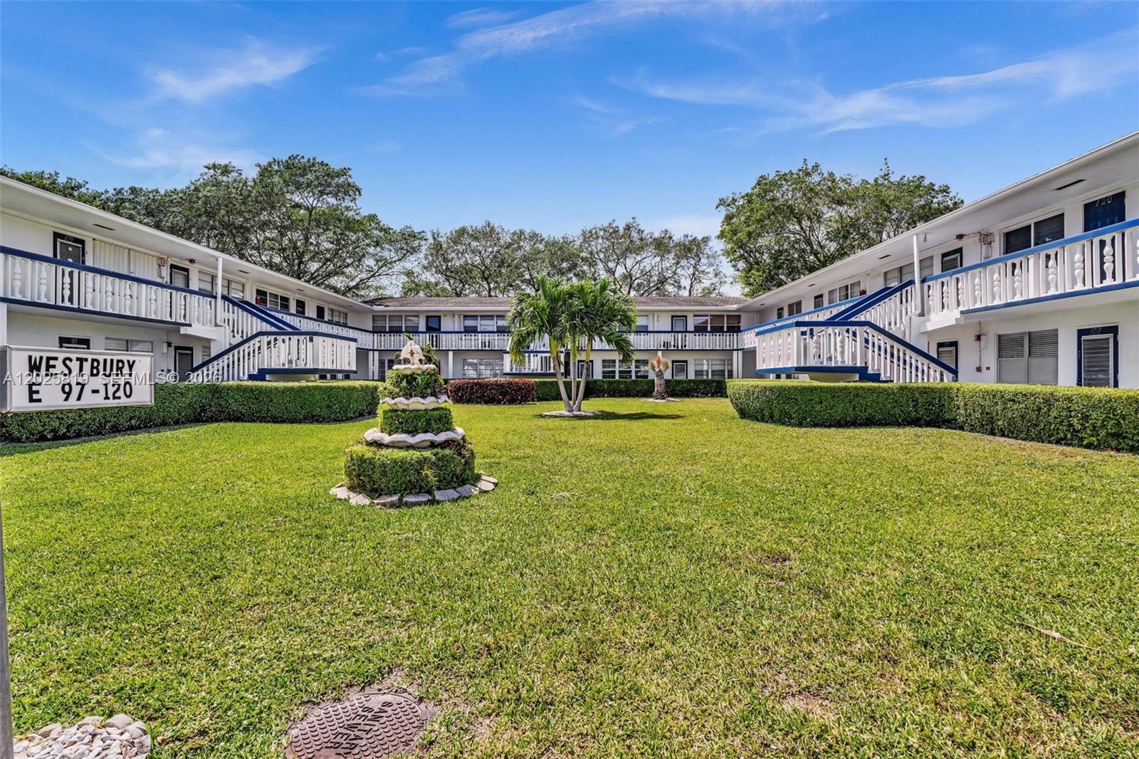 A two-story apartment complex surrounds a grassy courtyard with a tiered fountain and palm tree — 117 Westbury  E # 117, Deerfield Beach FL 33442, DEERFIELD BEACH, FL
