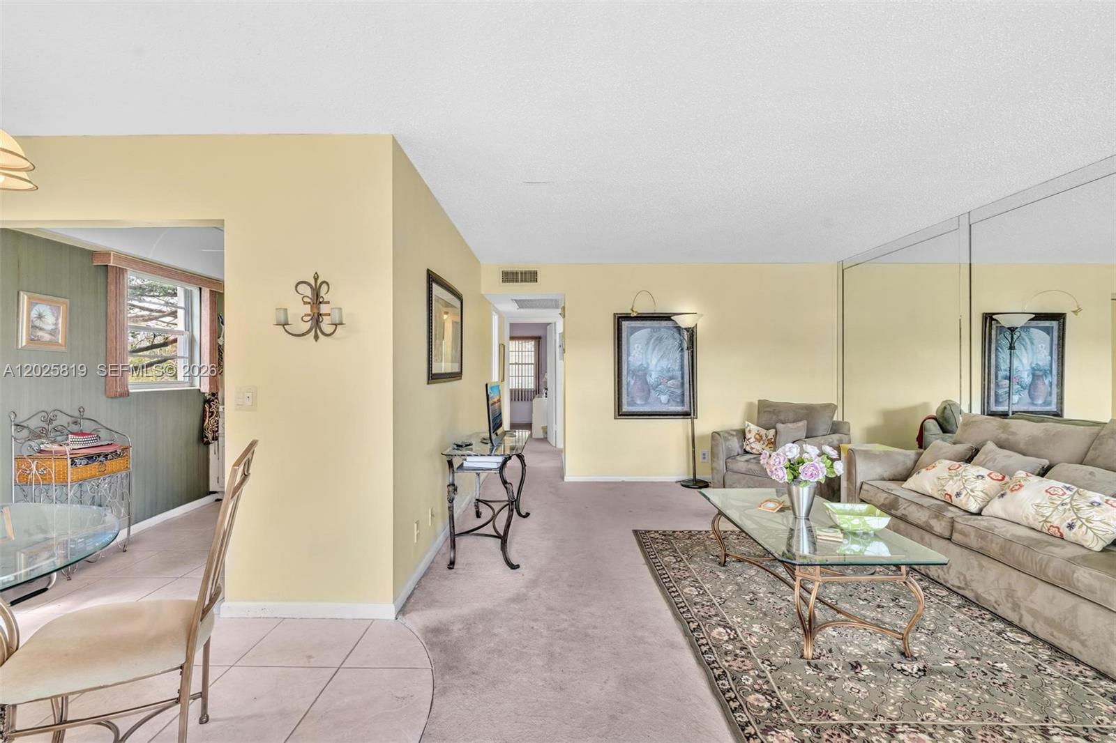 A living room features beige walls, carpeted flooring, a glass coffee table, and mirrored walls — 117 Westbury  E # 117, Deerfield Beach FL 33442, DEERFIELD BEACH, FL