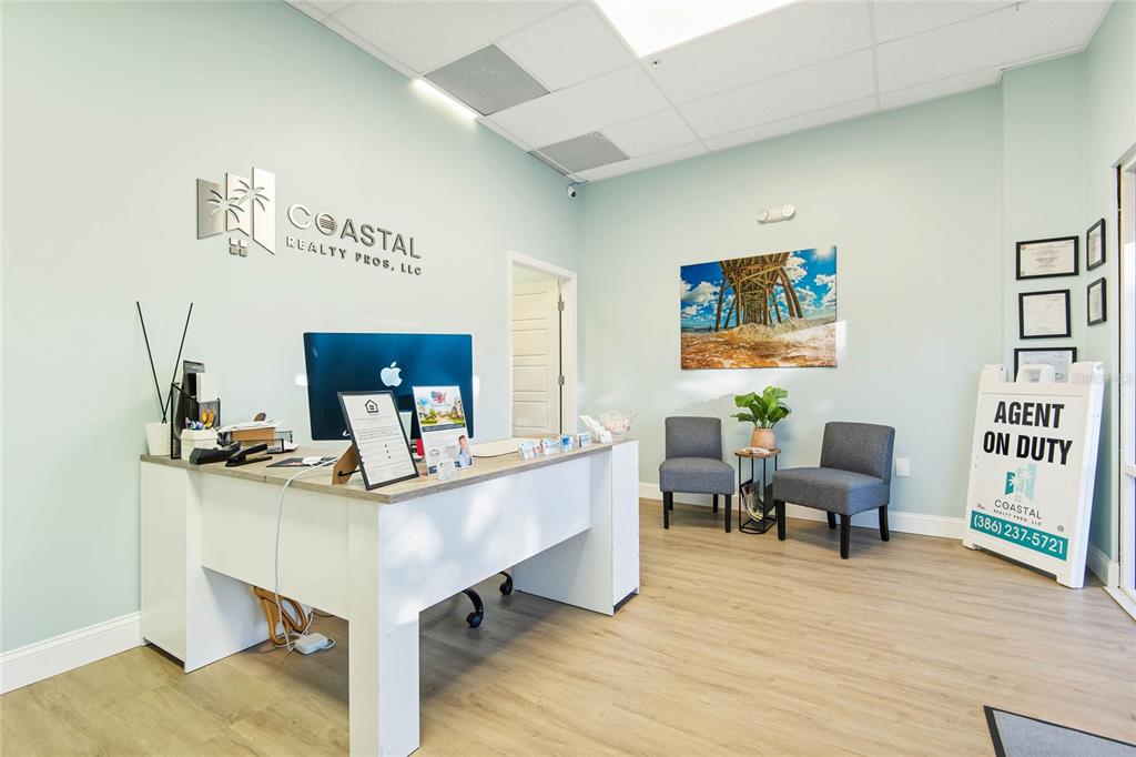 A real estate office features a white desk, two gray chairs, and a wall sign — 160 CYPRESS POINT PARKWAY UNIT C201 A, PALM COAST, FL