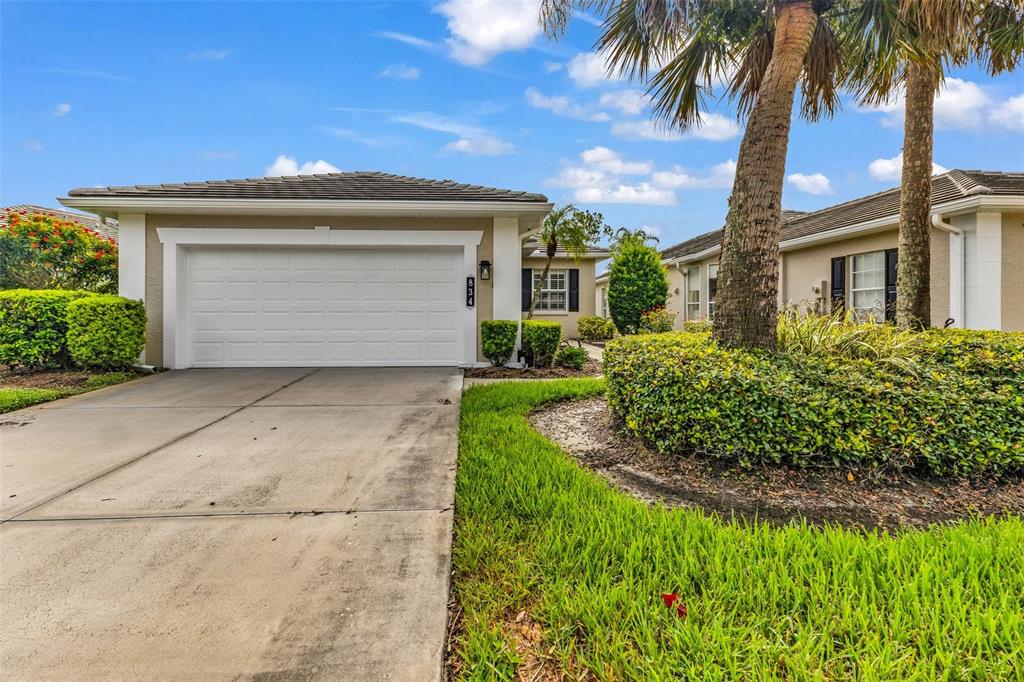 A single-story house exterior features a two-car garage, a concrete driveway, and landscaped greenery — 834 MONTROSE DRIVE UNIT 9, VENICE, FL