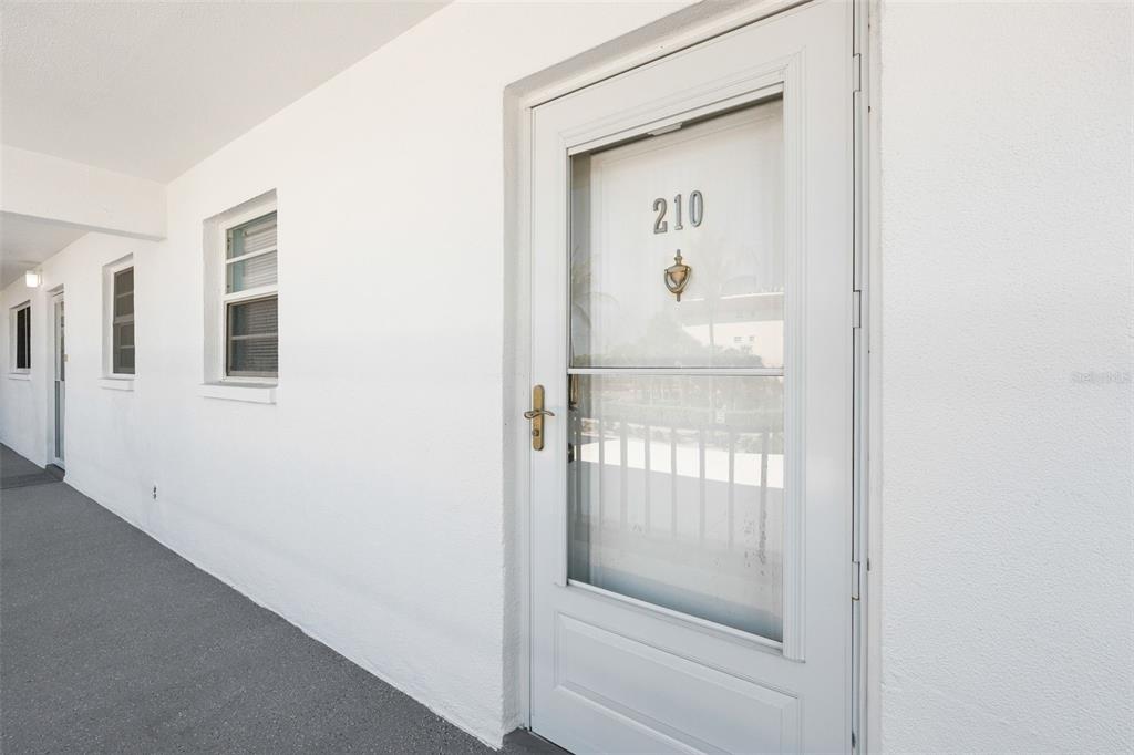 A white exterior door labeled 210 stands on a concrete walkway beside a building wall — 5600 BEACH WAY DRIVE UNIT 210, SARASOTA, FL